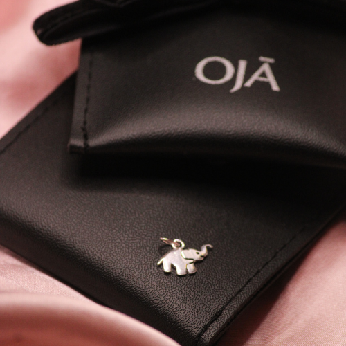Black leather wallet with 'OJá' branding and an elephant charm on a pink surface.